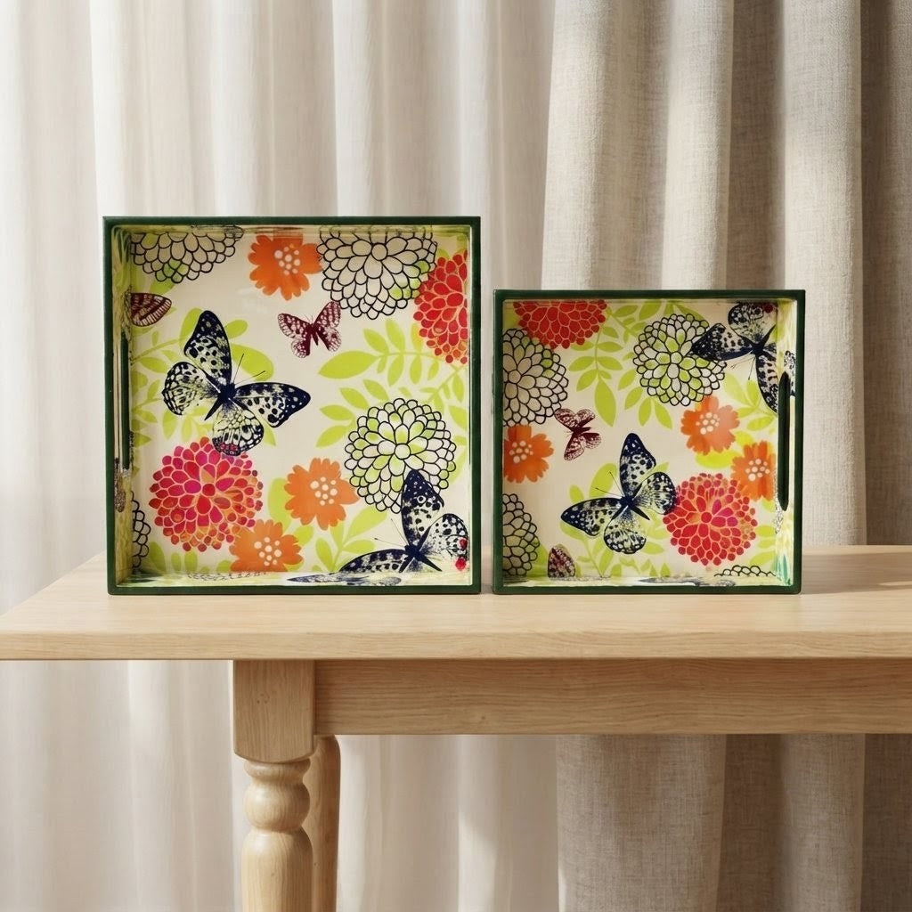 Two square decorative trays with a colorful floral and butterfly pattern, displayed side-by-side on a light wood table in front of beige curtains.