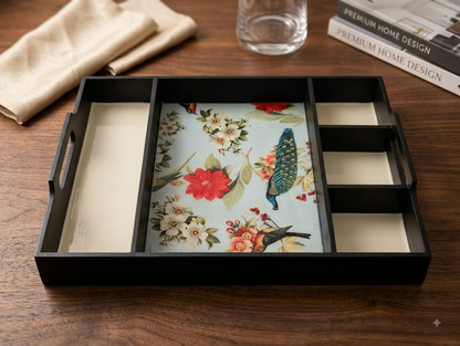 A black rectangular tray with divided compartments and handles, featuring a central floral and bird print insert, styled on a wooden tabletop with a folded beige cloth, a glass, and a magazine in the background.