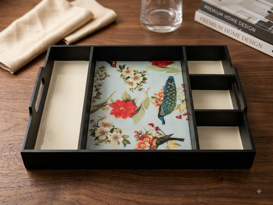 A black rectangular tray with divided compartments and handles, featuring a central floral and bird print insert, styled on a wooden tabletop with a folded beige cloth, a glass, and a magazine in the background.