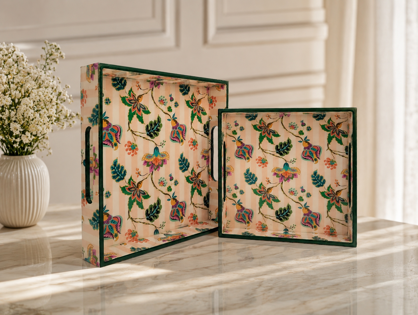 Two decorative serving trays with a colorful floral and bird pattern on a beige background, framed in dark green, displayed on a marble table next to a vase.