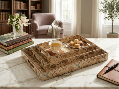 A set of three nested rectangular serving trays with a vintage floral and bird pattern in beige, brown, and muted blue, styled on a white marble table with a teacup, macarons, books, and a notebook in a bright, elegant room.