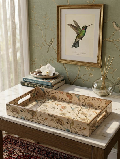A decorative rectangular serving tray with a floral and bird pattern sits on a marble-topped table in a styled room. A framed hummingbird print hangs on the wall above, with books, a white orchid, and a glass reed diffuser nearby.