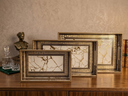 Three rectangular picture frames with ornate gold and bronze borders and marble-style inserts, arranged on a wooden table against a textured beige wall.