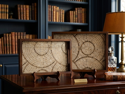 Two framed mosaic wall art pieces with circular geometric patterns in beige, brown, and cream tones, displayed on wooden stands on a dark wood desk in a library setting with bookshelves and a lamp in the background.