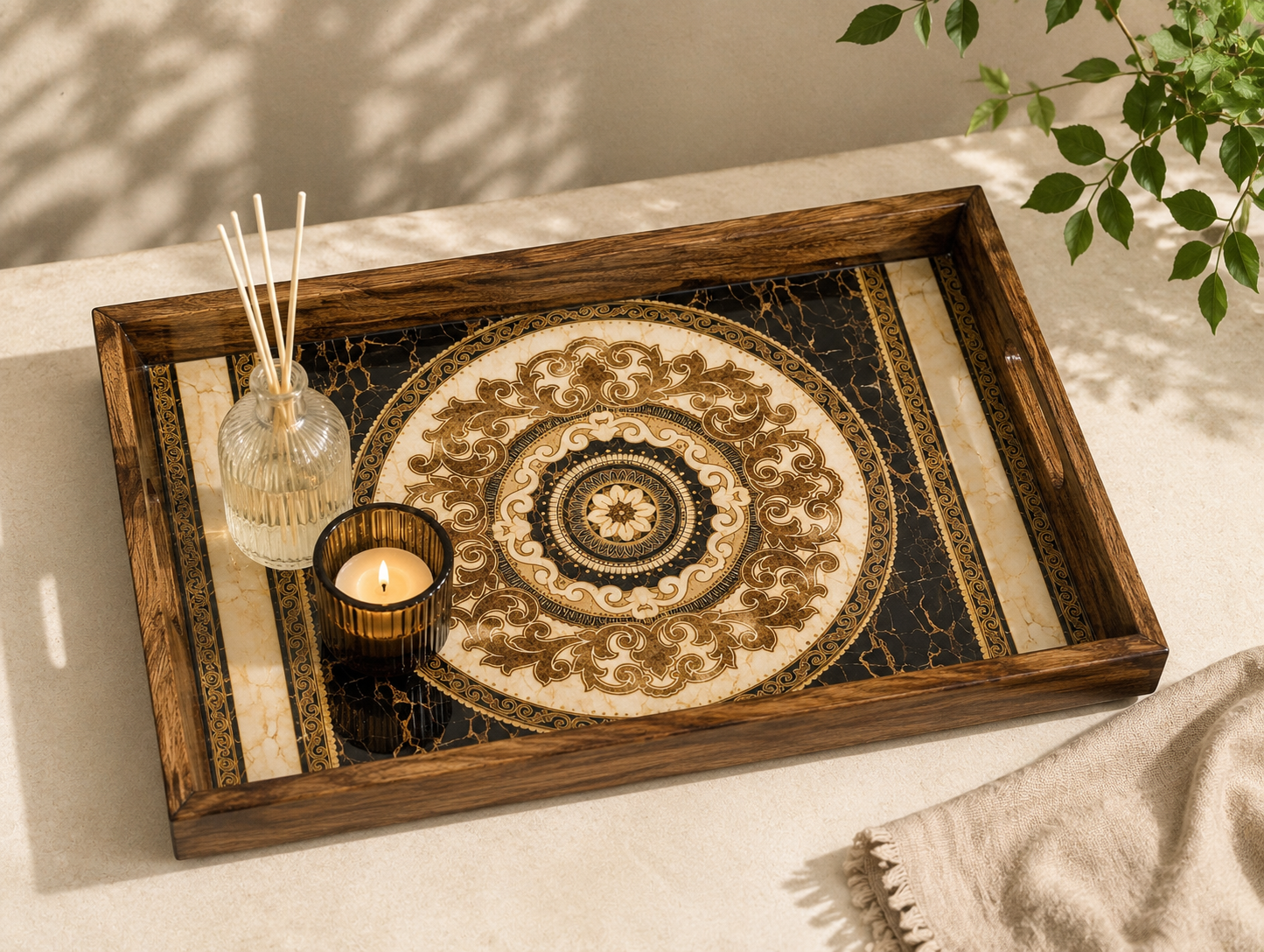 A rectangular wooden decorative tray with a raised rim, featuring an ornate circular medallion design in cream, gold, and black. The tray is styled on a light beige surface with a glass reed diffuser, a lit candle, and a beige throw blanket nearby.