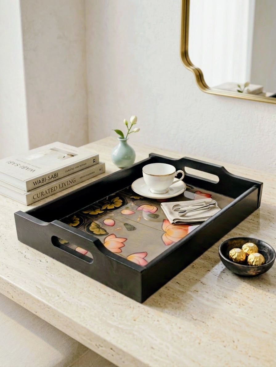A black rectangular serving tray with cutout handles sits on a light stone tabletop. The tray is styled with a floral-patterned liner, a white teacup and saucer, folded napkins, books, a small vase with a white flower, and a small bowl of chocolates.