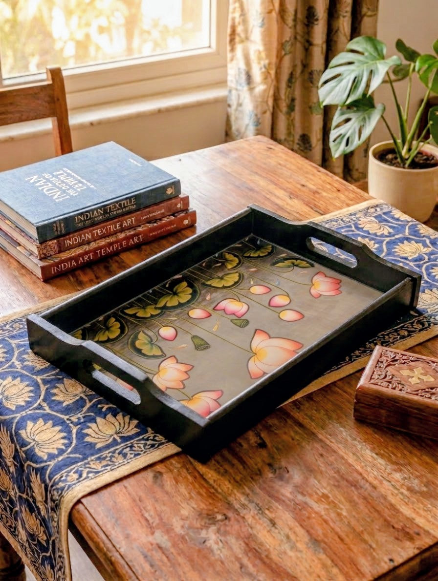 A rectangular black serving tray with floral hand-painted motifs in pink, orange, and green, displayed on a rustic wooden table next to books and a blue patterned runner.